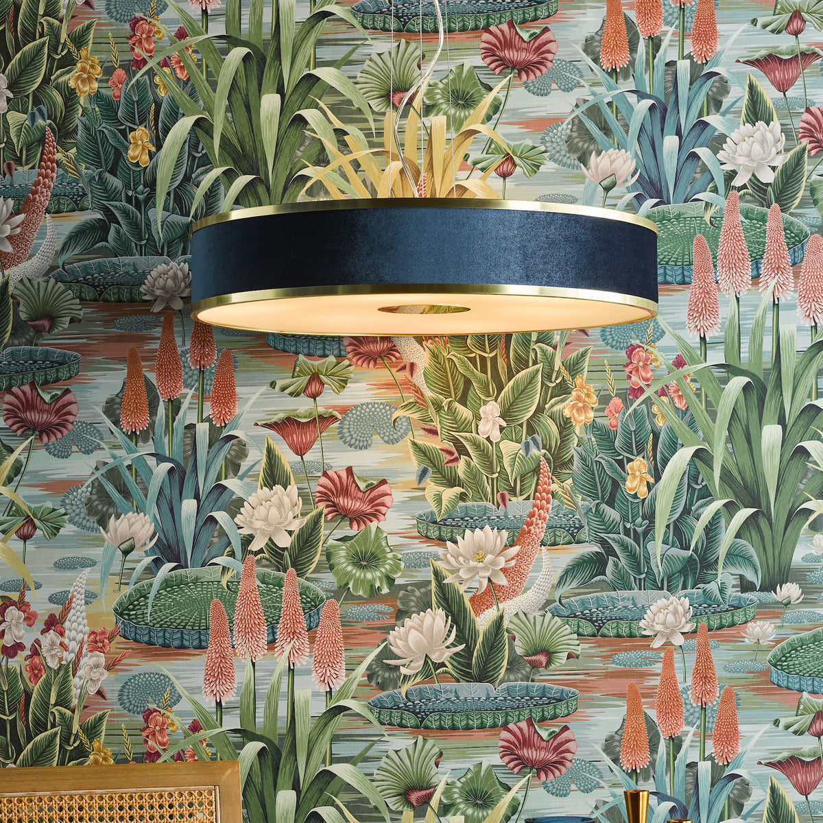 A blue and gold drum pendant light hangs in front of a botanical wallpaper with water lilies and red hot poker flowers.