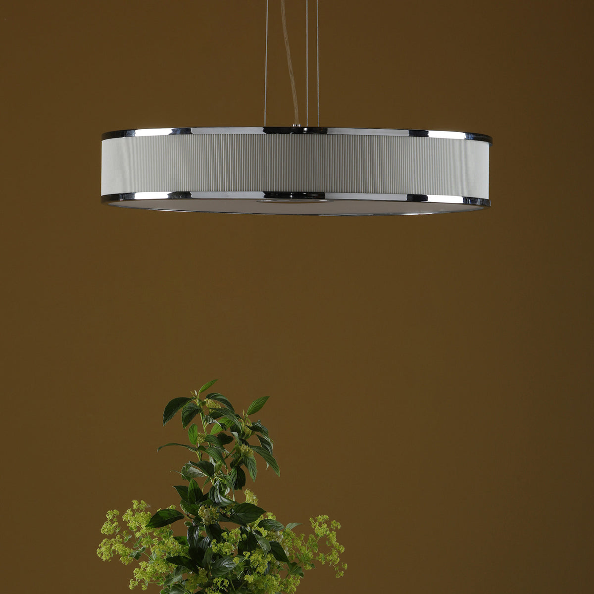 A modern, round pendant light with a white ribbed shade and chrome accents, hanging above green foliage.
