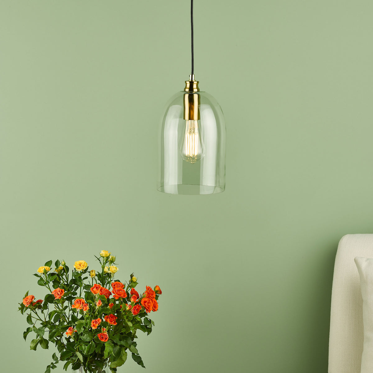 A clear glass pendant light with a brass fixture and an Edison bulb, hanging against a green wall.
