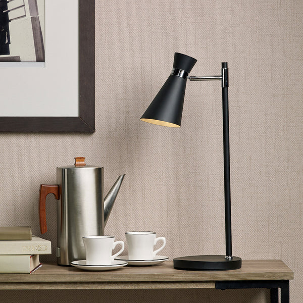 A black desk lamp with a conical shade illuminates a wooden table with a silver coffee pot and two white cups.
