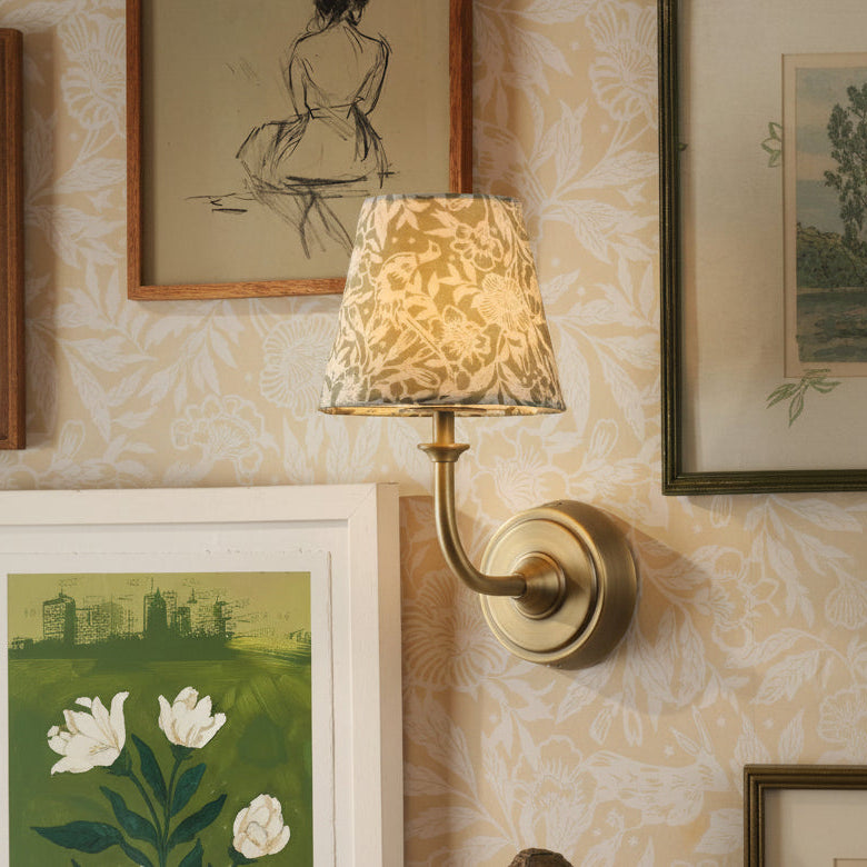 Brass wall sconce with a patterned fabric shade mounted on floral wallpaper