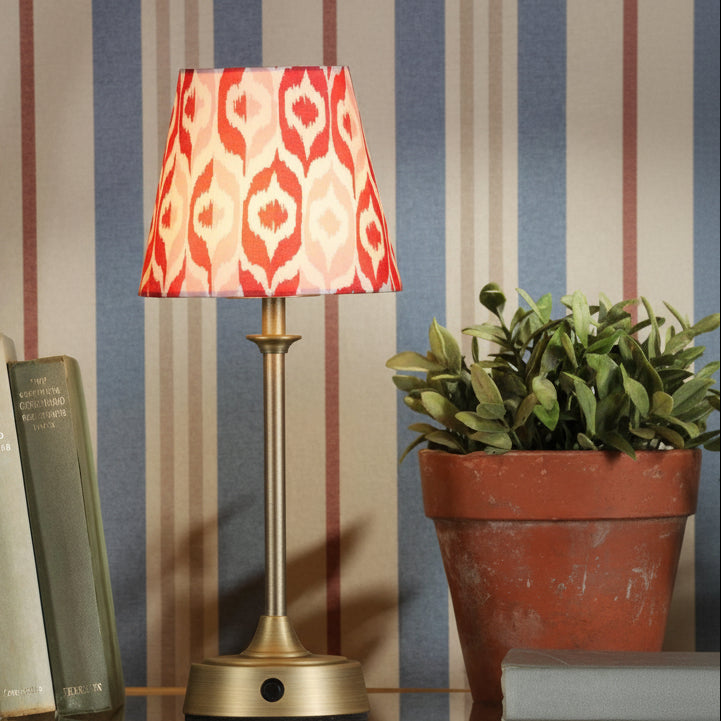 Table lamp with a red and white patterned shade on a brass base next to a potted plant and books