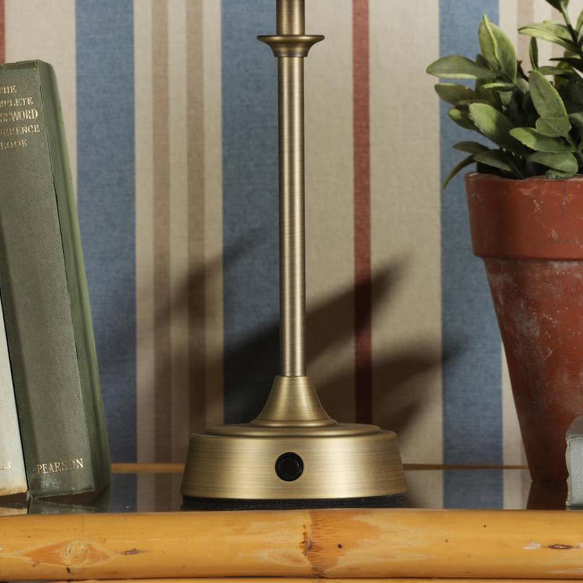 Antique brass lamp base with a push button switch on a wooden shelf next to a book and a potted plant