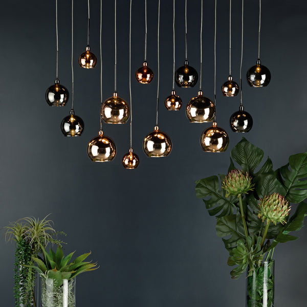Multiple pendant lights with round, reflective shades hang at varying heights against a dark wall. Green plants are below.