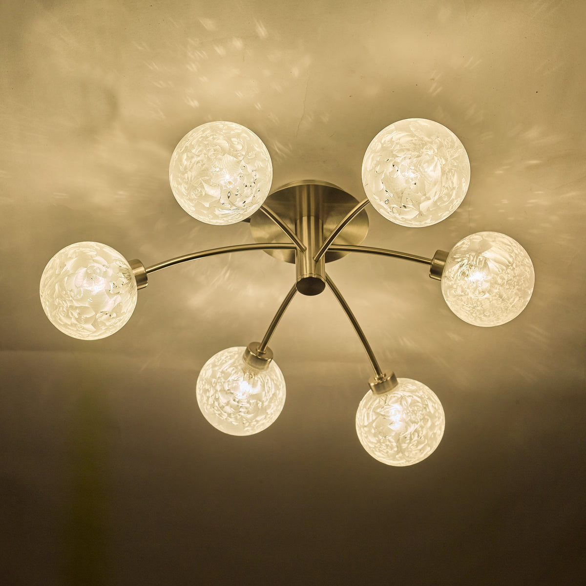 Modern ceiling light with six frosted glass globes on curved brass arms, casting warm light and patterns.
