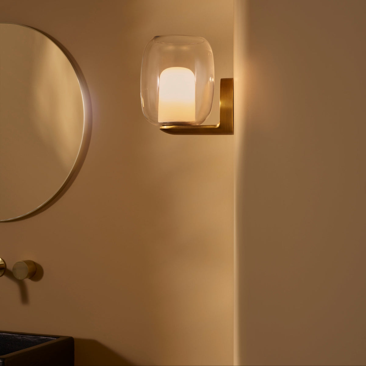 Wall-mounted light fixture with a clear glass shade and a brass base, next to a round mirror