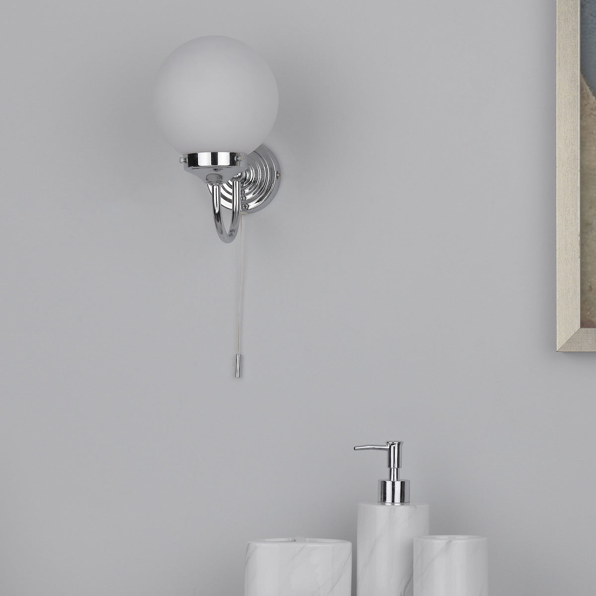 Chrome wall light with a frosted globe shade and a pull cord, above marble bathroom accessories.
