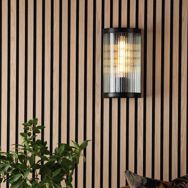 A black and wood slatted wall with a cylindrical glass wall light and green plant