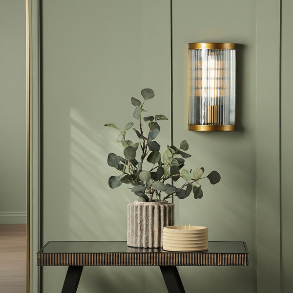 Gold and glass wall sconce above a console table with a plant in a concrete pot and a yellow candle