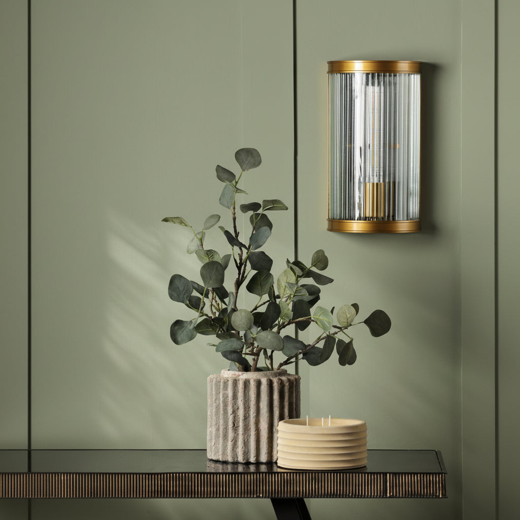 A gold and glass wall light above a plant in a textured pot and a yellow candle on a dark console table