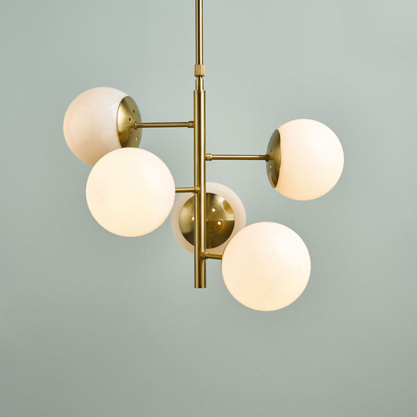 Modern brass chandelier with five glowing white globe lights against a light blue wall.