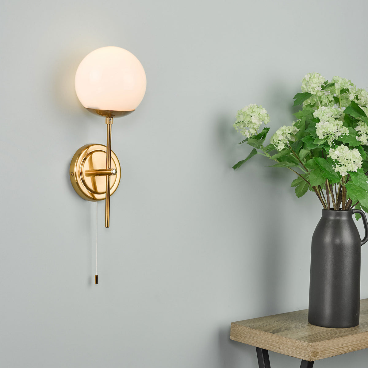 Brass wall light with a white globe shade and pull chain, next to a black vase with green flowers