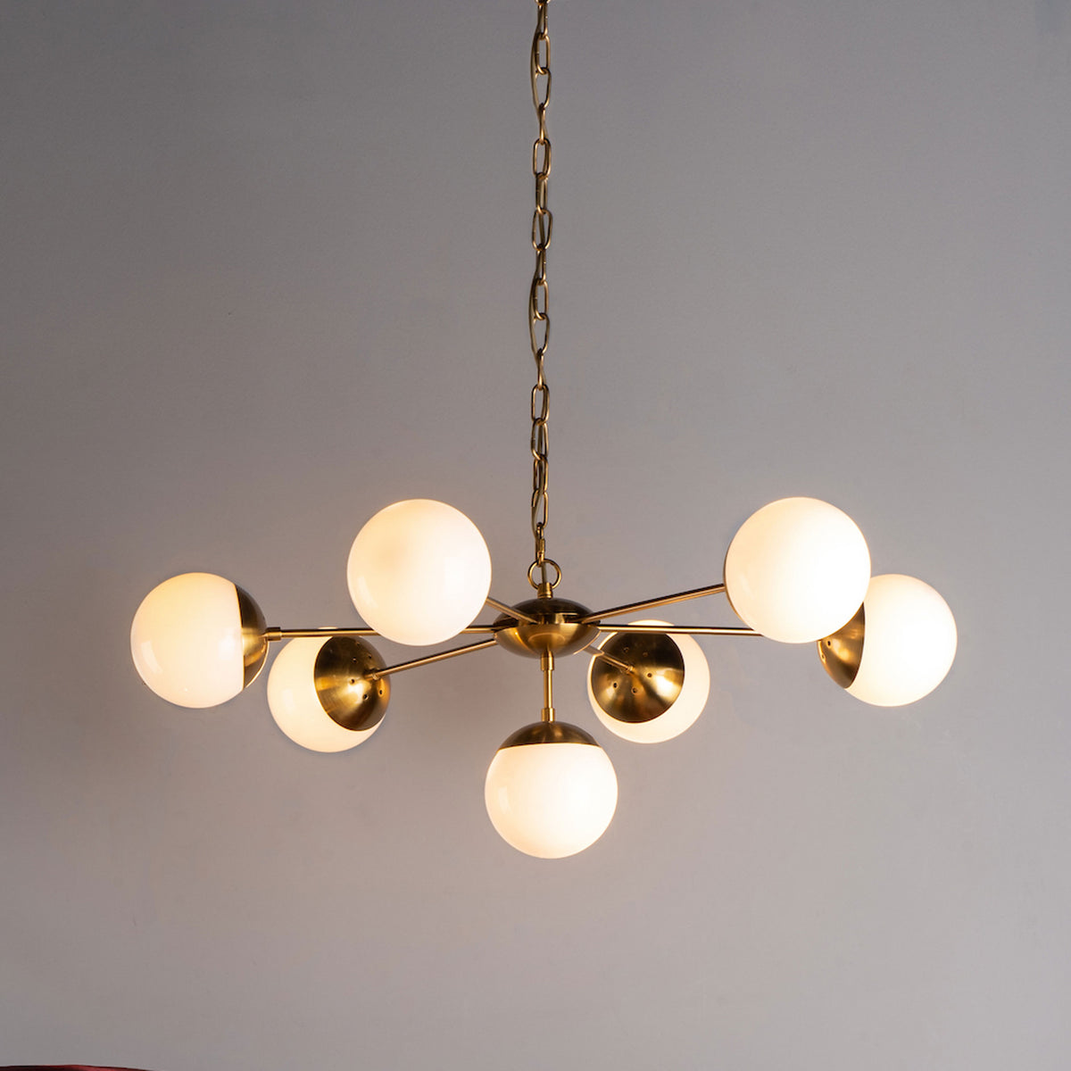 Modern brass chandelier with seven white globe lights, hanging from a chain against a gray wall.