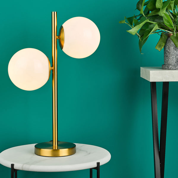 Gold table lamp with two spherical white lights on a marble table against a teal wall