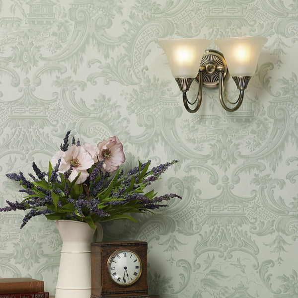 A wall sconce with two lights illuminates a wall covered in light green patterned wallpaper.