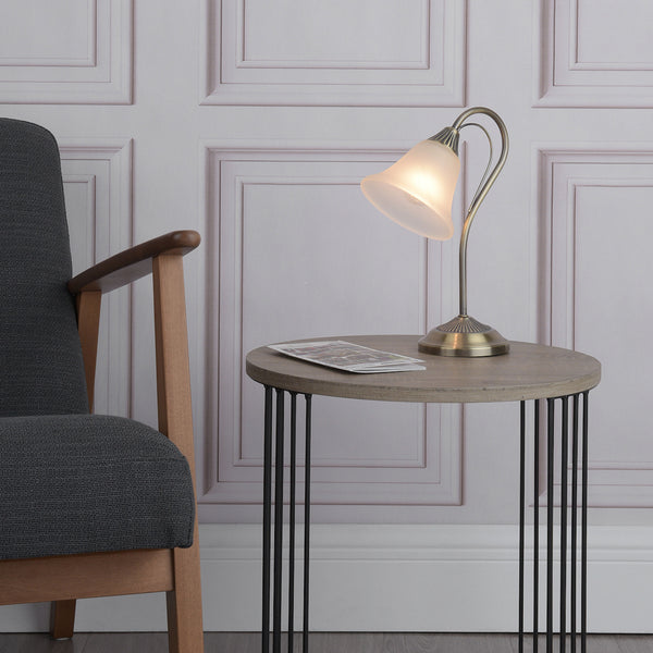 A lit table lamp with a frosted glass shade on a round wooden table next to a gray armchair.