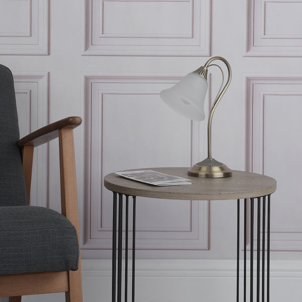 A bronze table lamp with a frosted glass shade sits on a round wooden table next to a gray armchair.