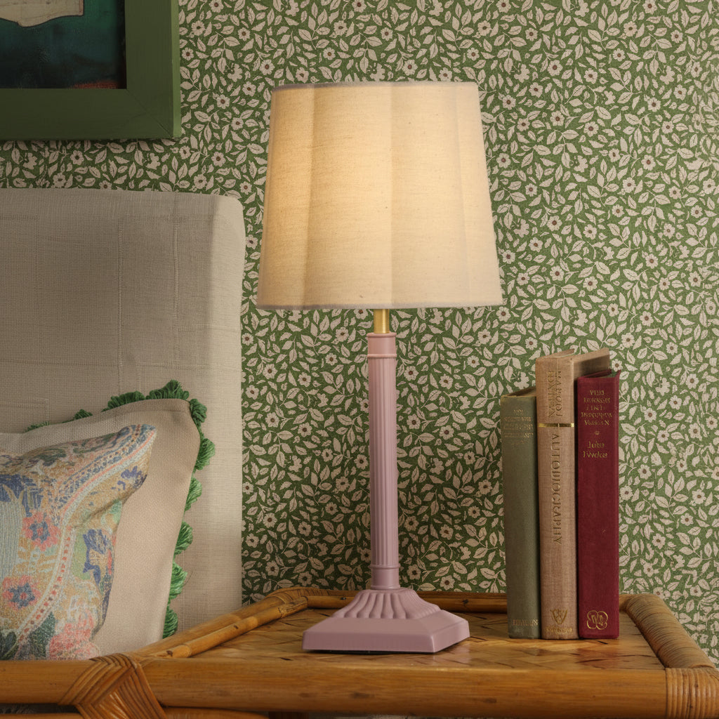Pink table lamp with beige fabric shade on a rattan nightstand next to books