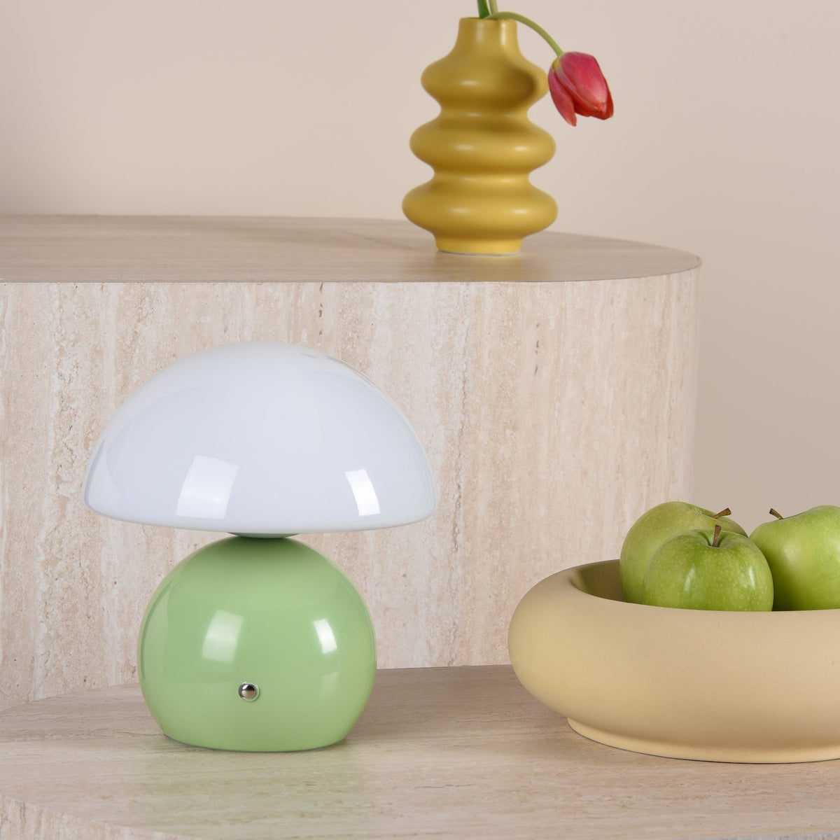 Mushroom lamp with a green base and white top, next to a yellow vase with a red tulip and a bowl of green apples