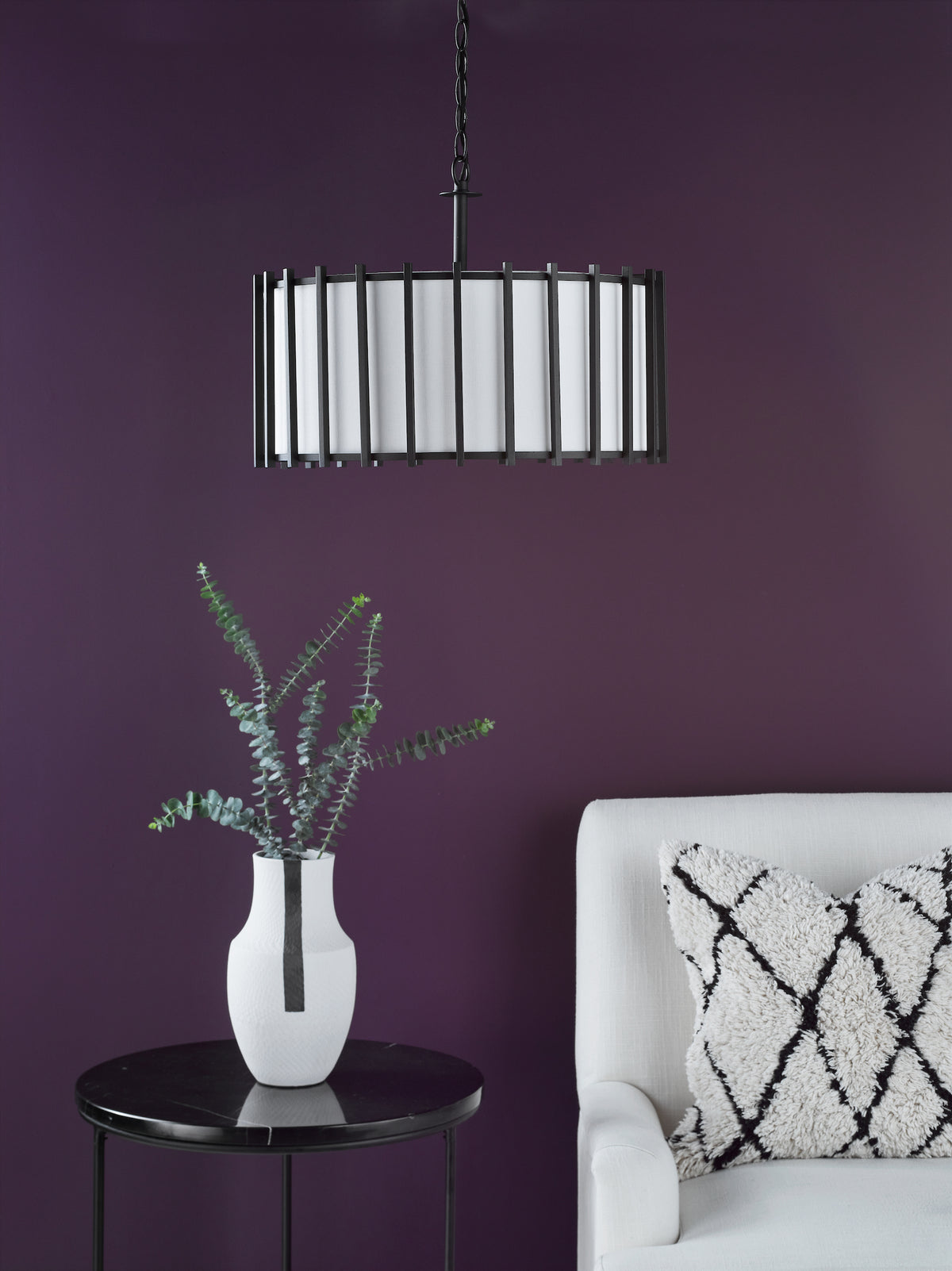 Black pendant light with white shade, black side table with white vase and eucalyptus, white chair with pillow