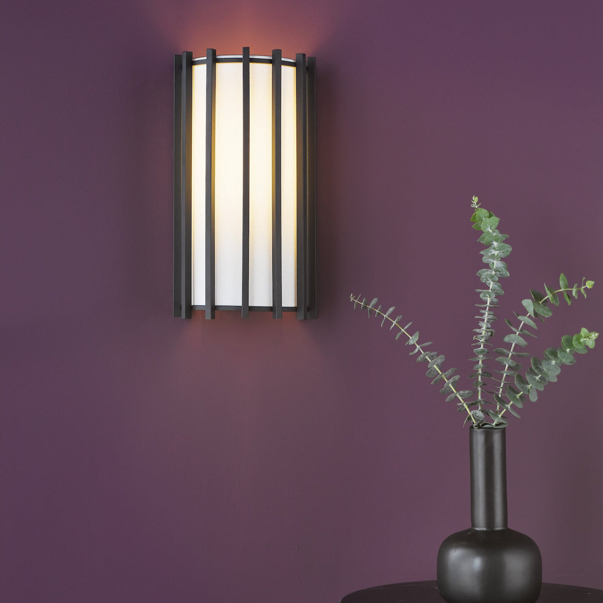 Wall light with a black frame and white shade, next to a black vase with eucalyptus branches