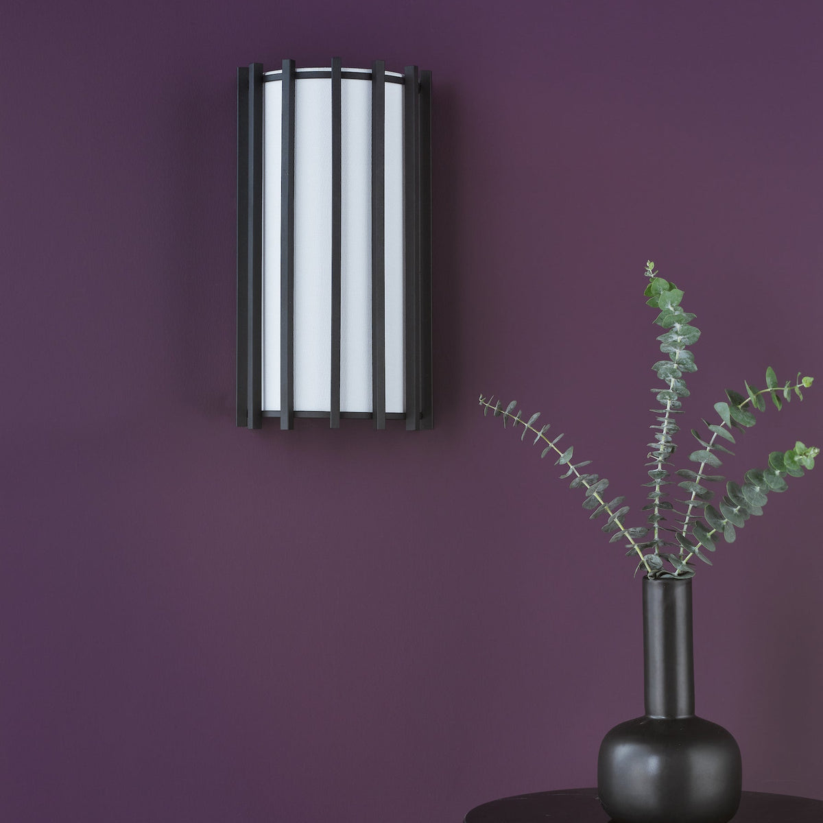 Wall sconce with black frame and white shade, next to a black vase with eucalyptus branches