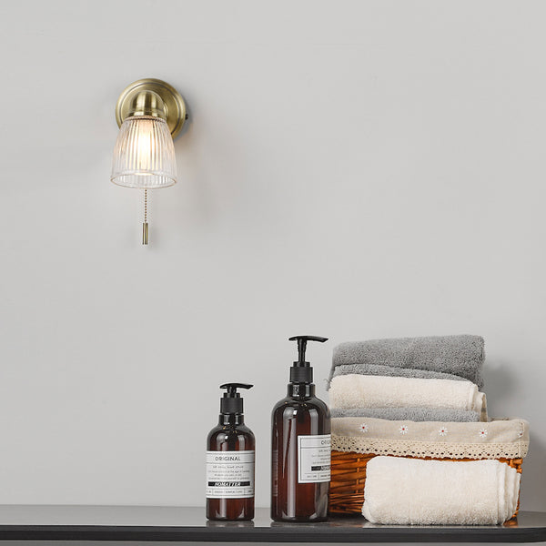 A brass wall sconce with a ribbed glass shade and pull chain, above two amber pump bottles and a basket of towels.