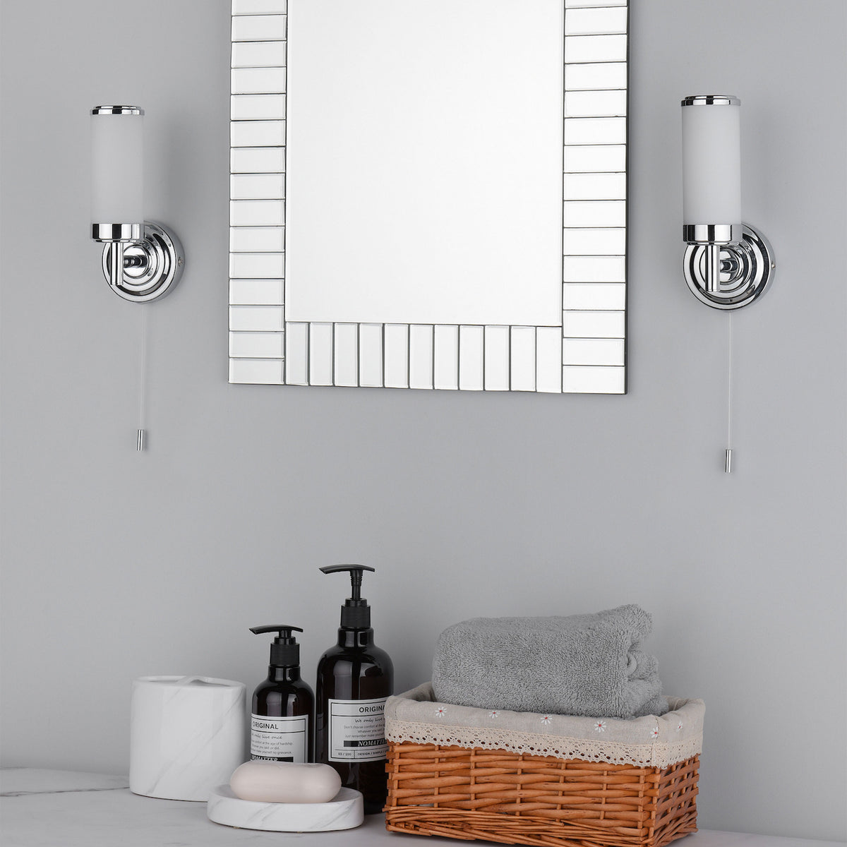 Bathroom vanity with two chrome wall sconces, a mirror, soap dispensers, a soap dish, and a towel in a basket.