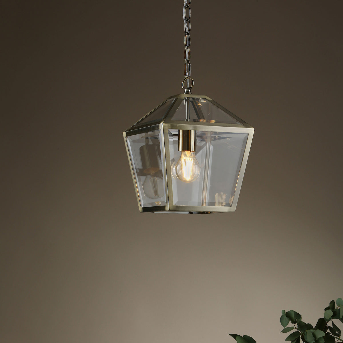 A brass and glass lantern-style pendant light with an exposed filament bulb, hanging against a warm brown wall.