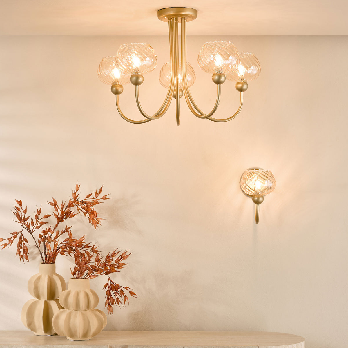 Gold chandelier with five glass shades and a matching wall sconce above two vases with dried foliage