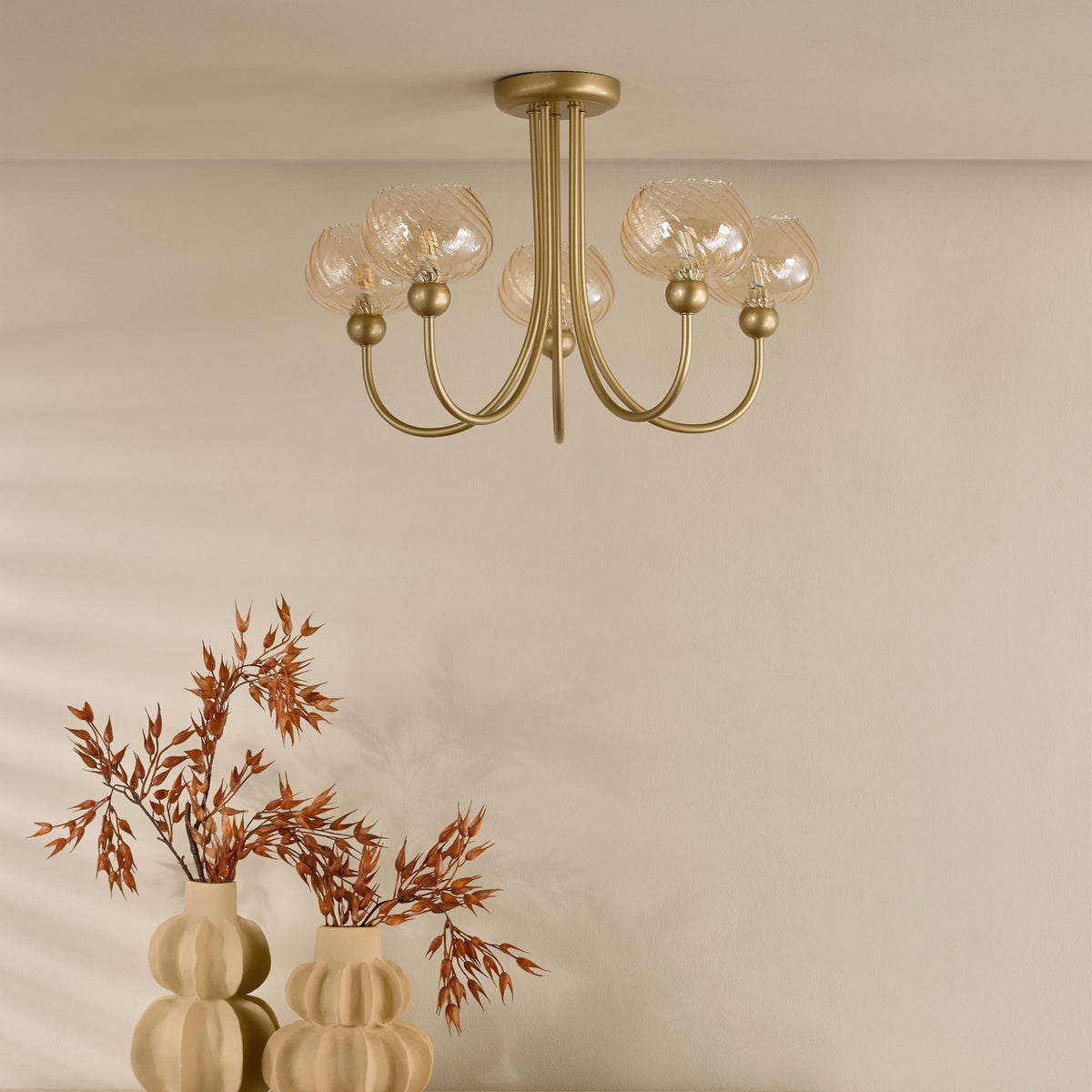Gold chandelier with five amber glass shades, two beige vases with dried orange foliage on a beige wall