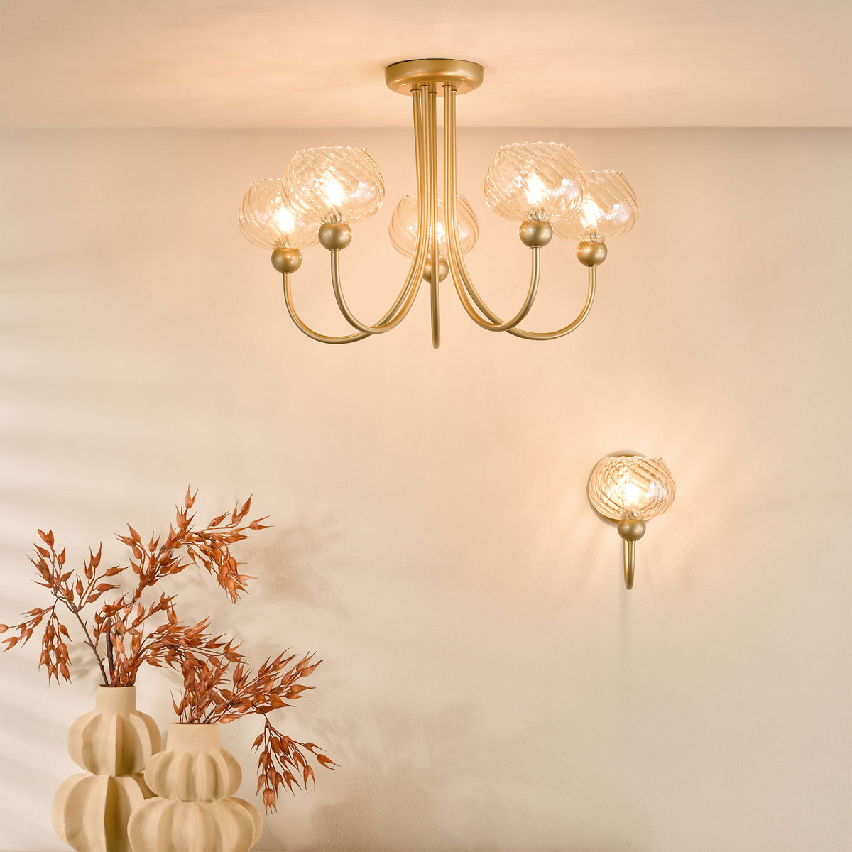 Gold chandelier with five glass shades and a matching wall sconce, above two cream vases with dried plants