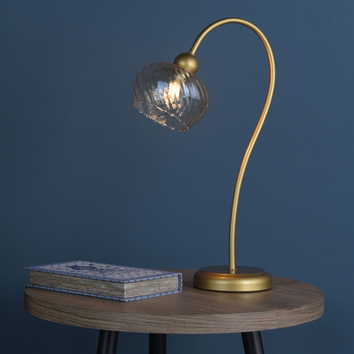 Gold table lamp with a clear glass shade and a book with a blue and white cover on a wooden table
