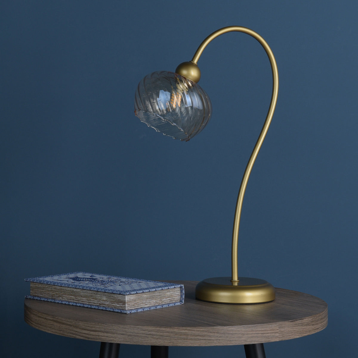 Gold table lamp with a clear, textured glass shade next to a blue patterned book on a wooden table
