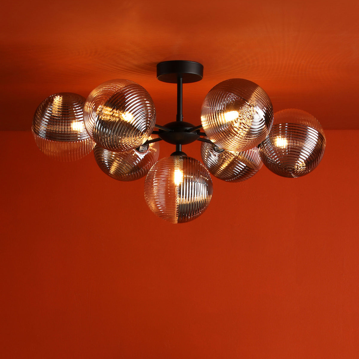 Modern black chandelier with seven ribbed glass globes, illuminated against a vibrant orange ceiling and wall.