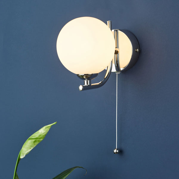 A chrome wall light with a spherical opal glass shade and a pull cord switch, against a dark blue wall.