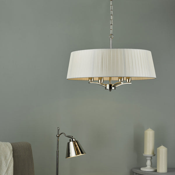 A white pleated drum chandelier hangs above a silver floor lamp and two white candles.