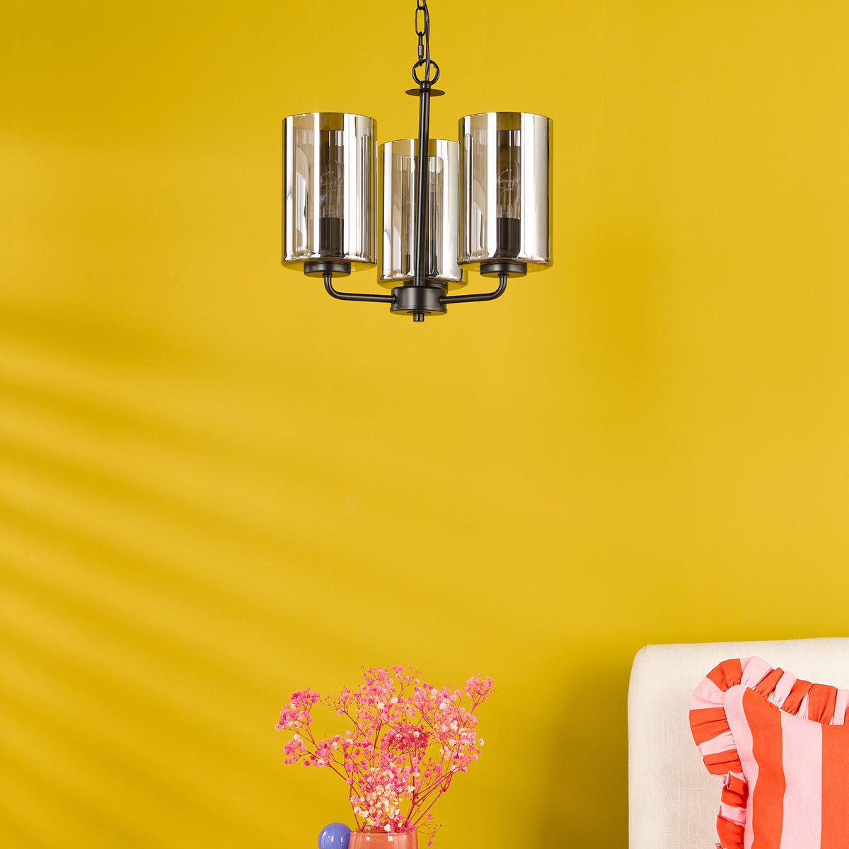 Black metal chandelier with three clear glass shades, pink flowers in a vase, and a striped pillow