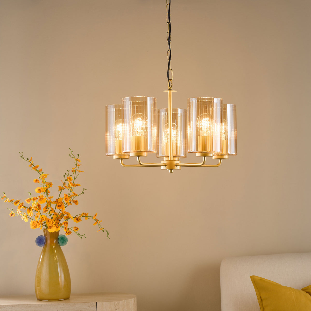 Gold chandelier with six glass shades and exposed bulbs, illuminating a room with a vase of yellow flowers