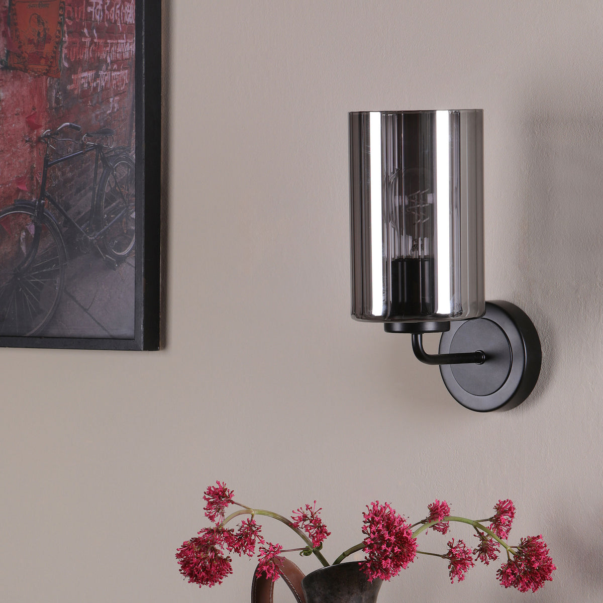 Black wall sconce with a smoked glass shade, next to a framed bicycle print and pink flowers