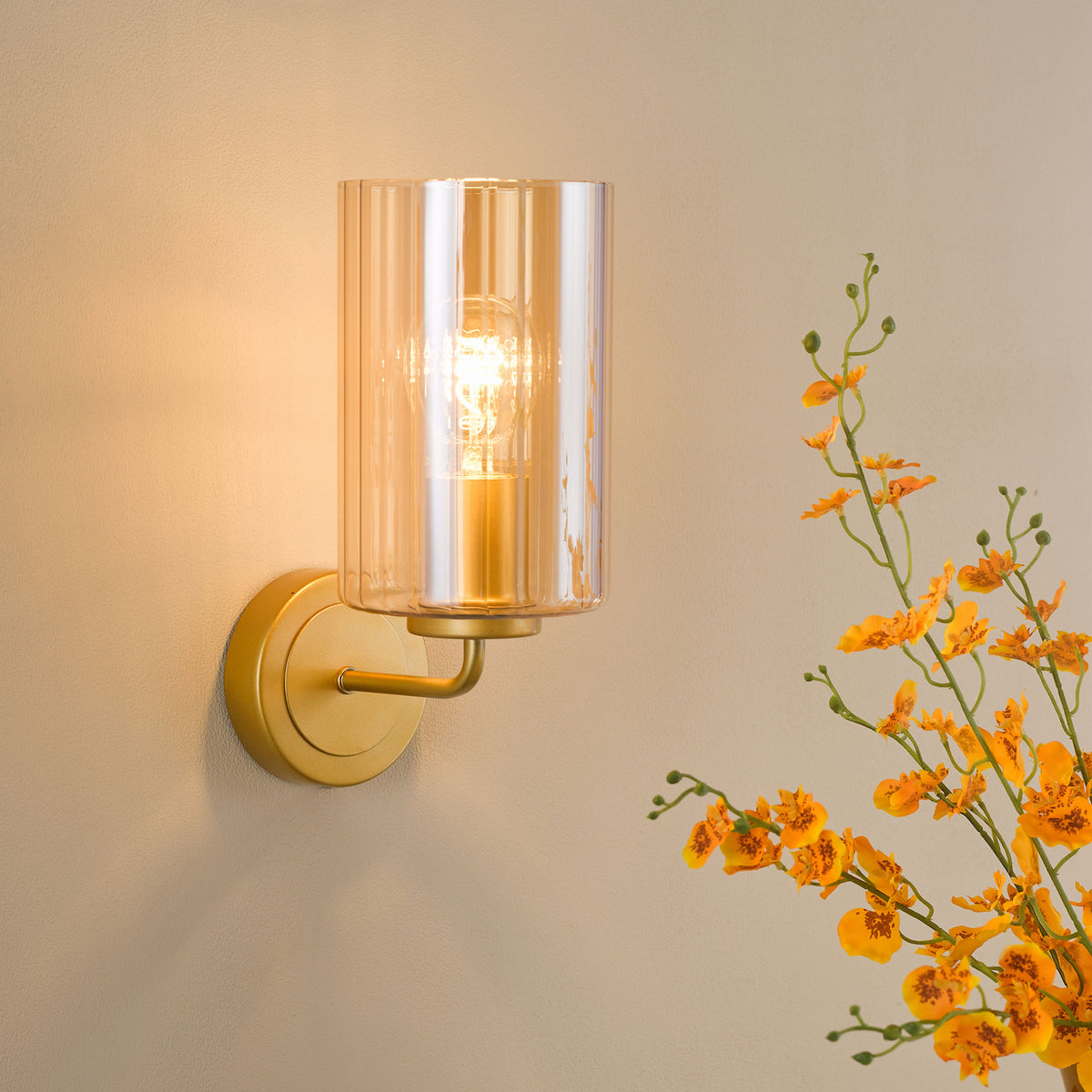 Gold wall sconce with a ribbed amber glass shade and exposed bulb, next to orange flowers