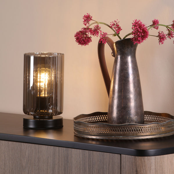 A table lamp with a smoked glass shade and a metal pitcher with pink flowers on a wooden cabinet