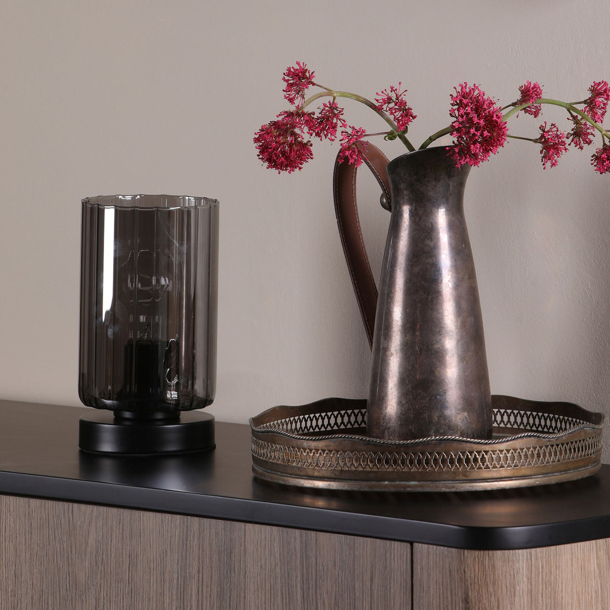 Table lamp with ribbed glass shade next to a metal pitcher with pink flowers on a wooden cabinet