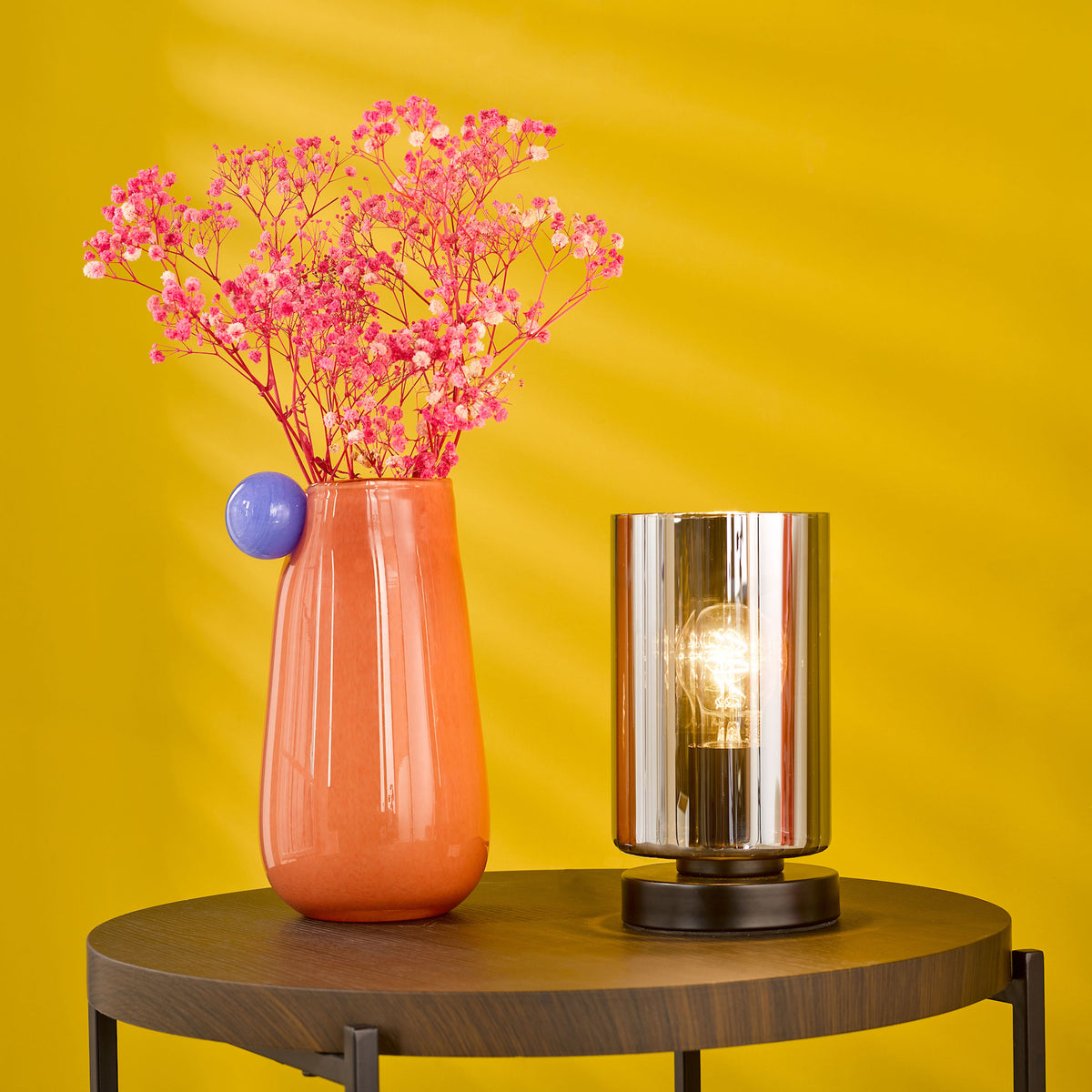 Orange vase with pink flowers and a lit table lamp on a round wooden table