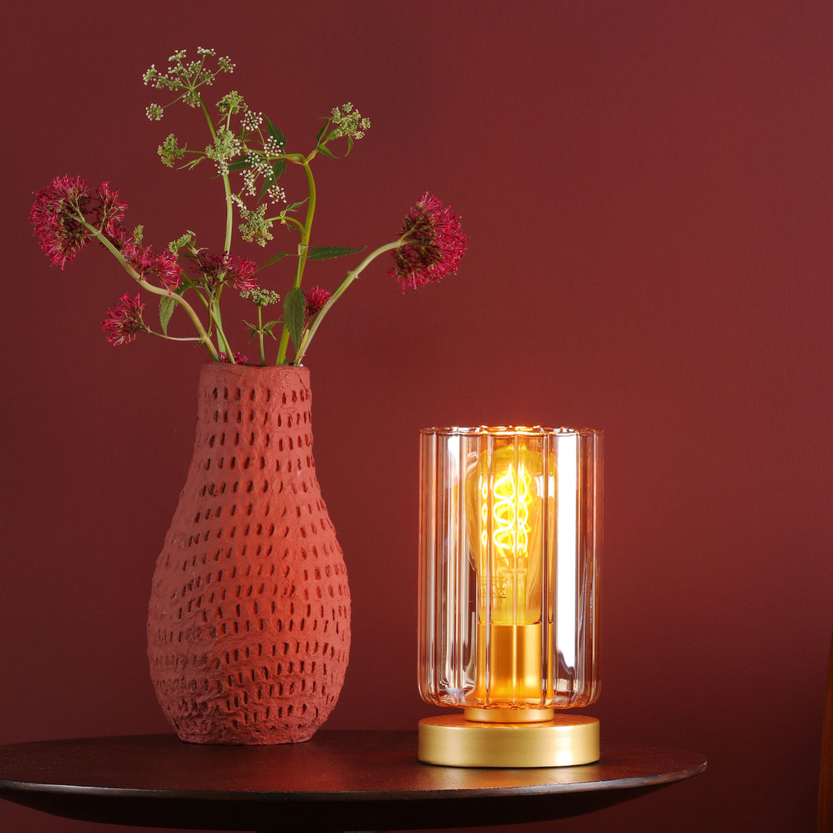 Red vase with flowers next to a gold and glass lamp on a dark table