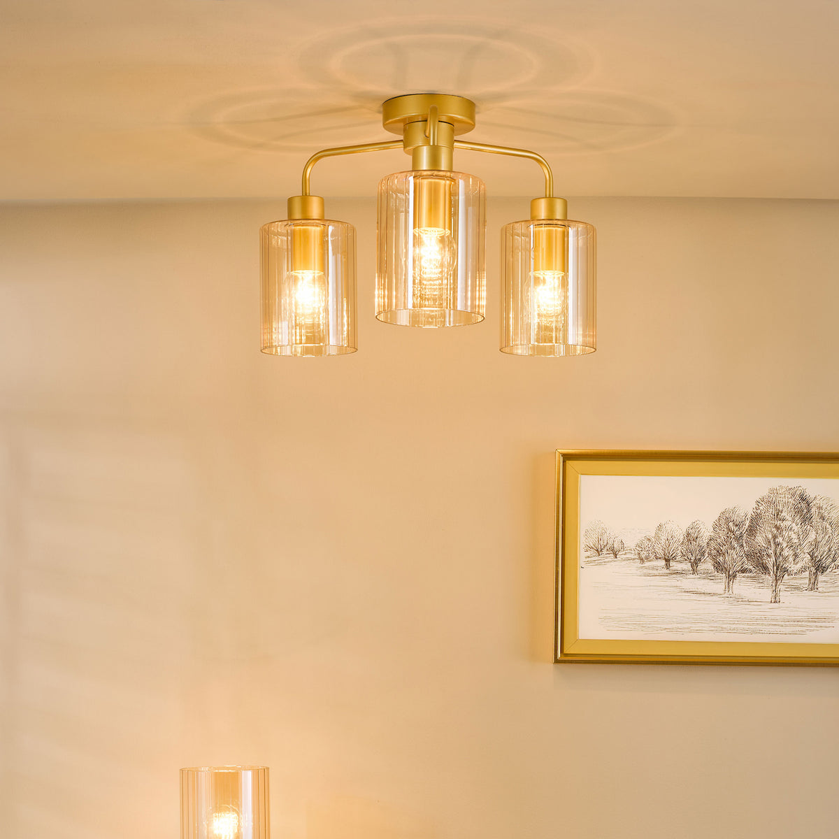 Gold ceiling light with three ribbed glass shades, illuminating a wall with a framed tree drawing