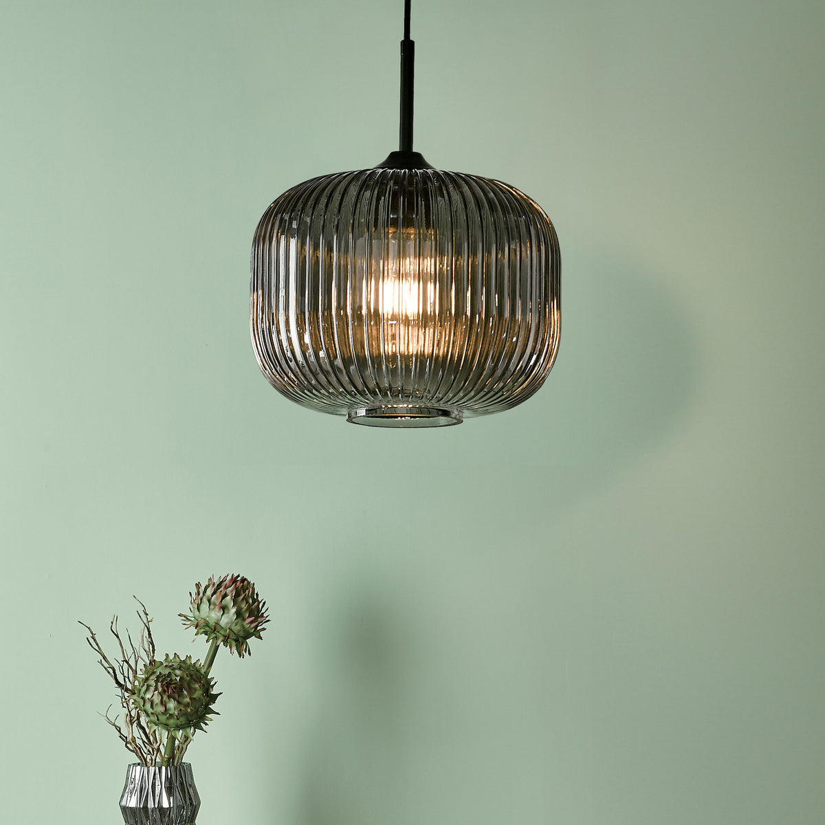 A ribbed glass pendant light hangs above a vase with dried branches and two green artichoke flowers.