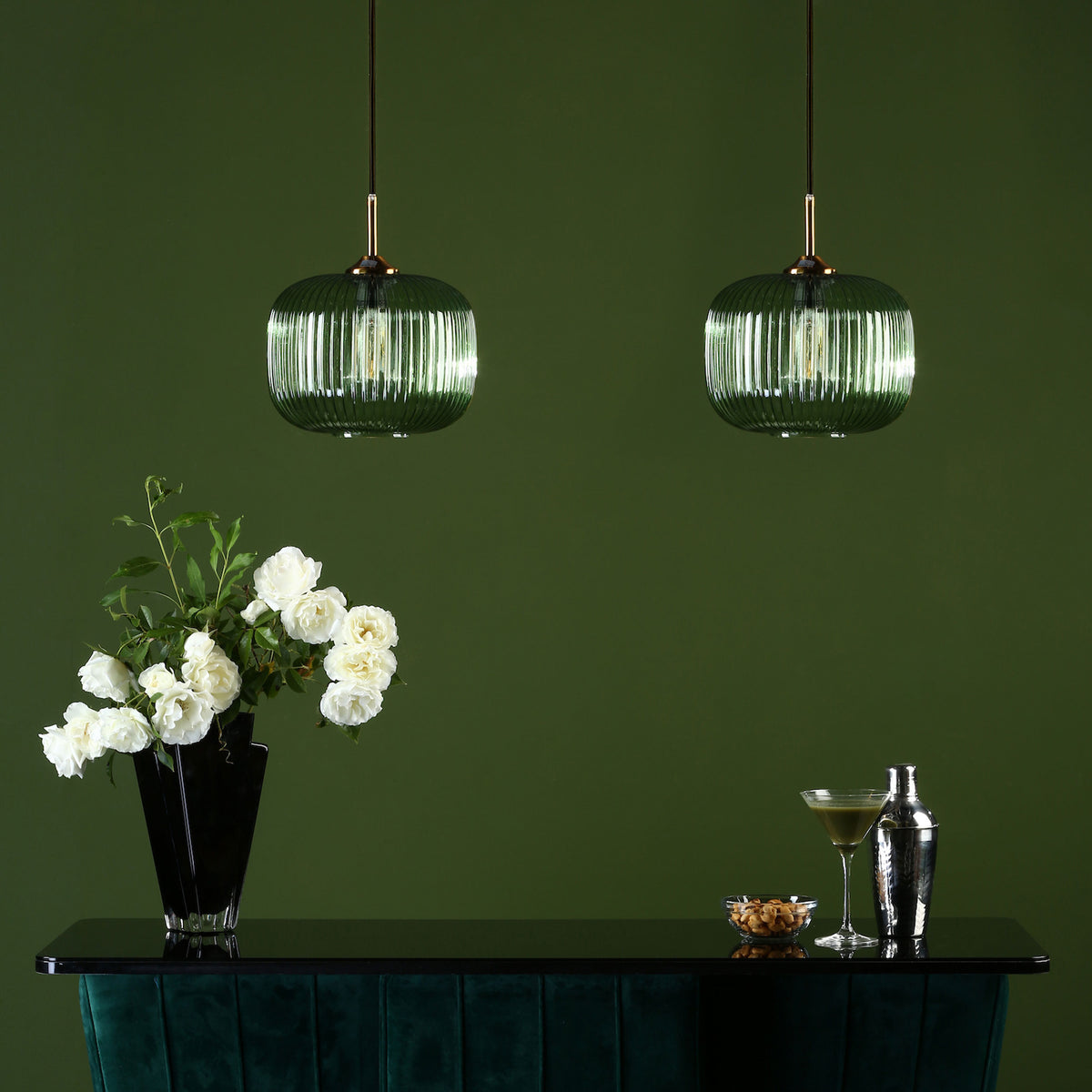 Two green ribbed glass pendant lights illuminate a bar with white roses, a cocktail, and nuts.