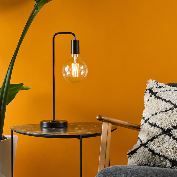 Black table lamp with a clear bulb on a marble-top side table next to a plant and a textured pillow.