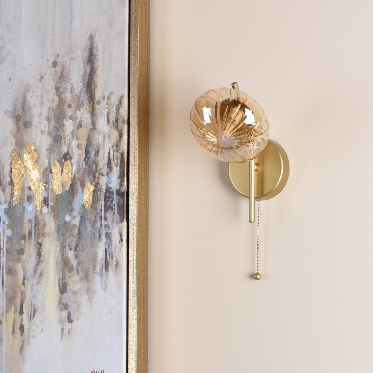 Gold wall sconce with a textured amber glass shade and a pull chain next to a framed abstract painting